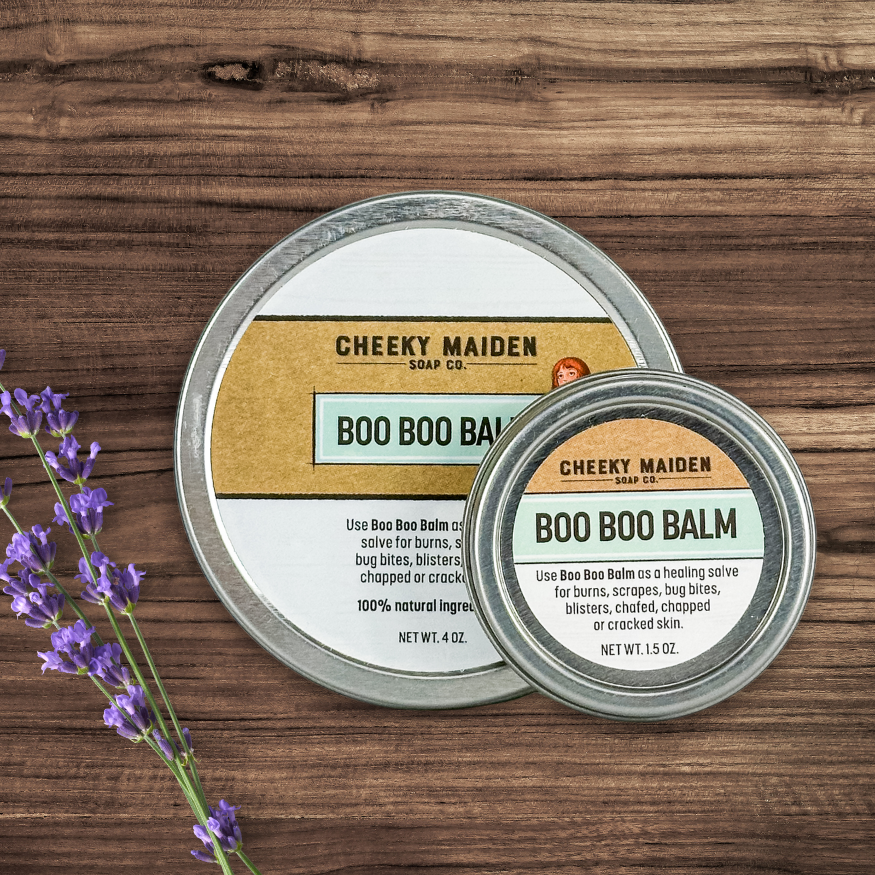 Two tins of Cheeky Maiden Soap Co. Boo Boo Balm on a wooden surface with lavender.