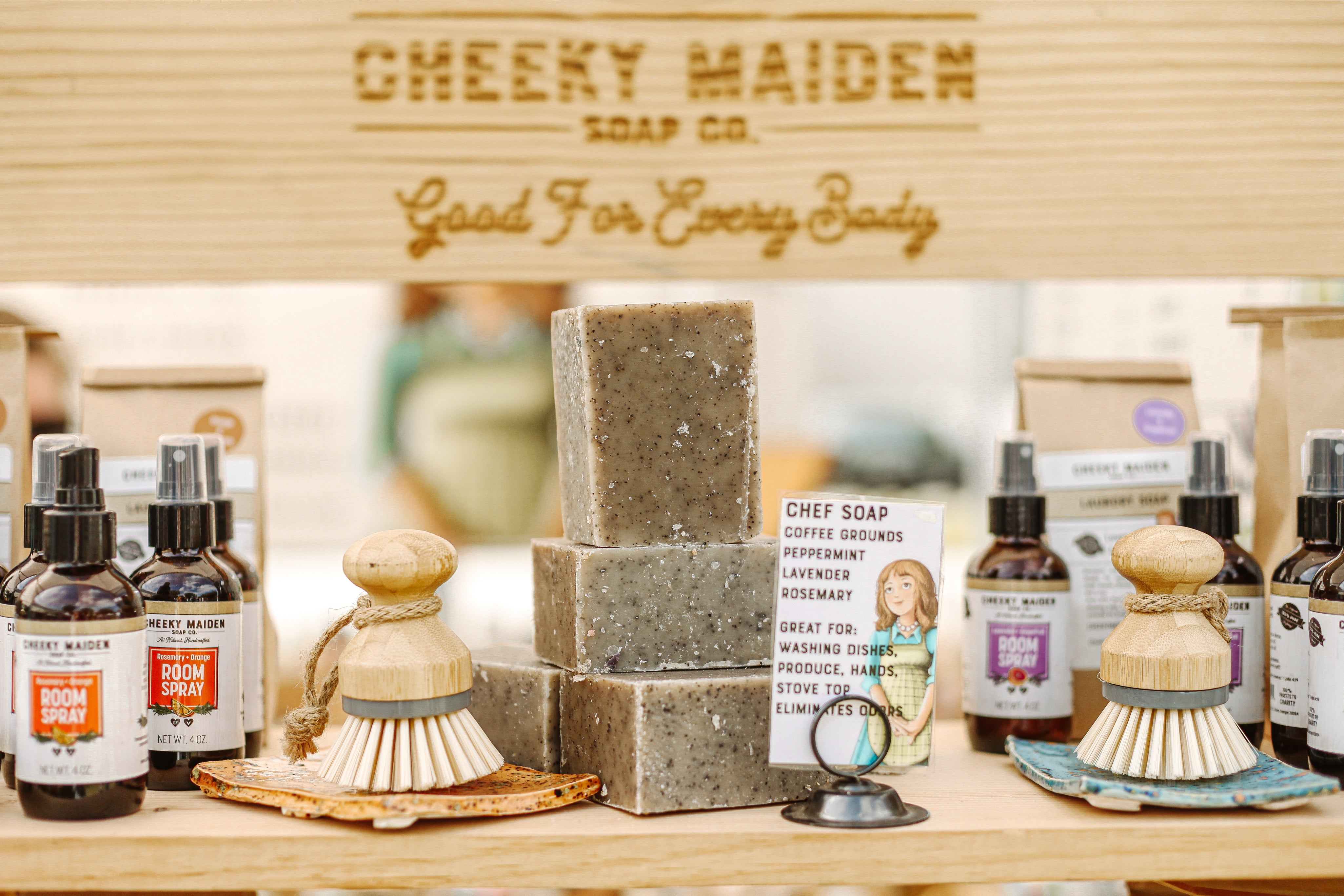 Cheeky Maiden Soap Co products including soap bars, bottles, and a brush on a wooden surface.