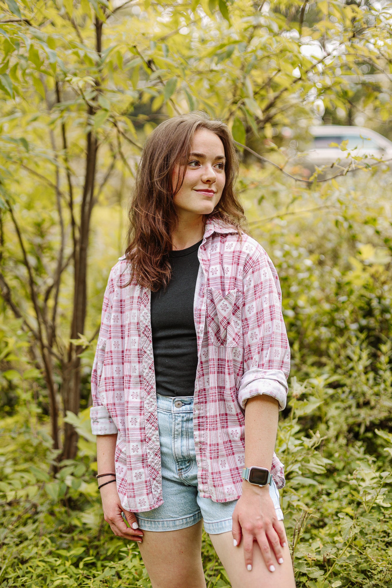 Kayla standing in a forest wearing a plaid shirt and denim shorts.