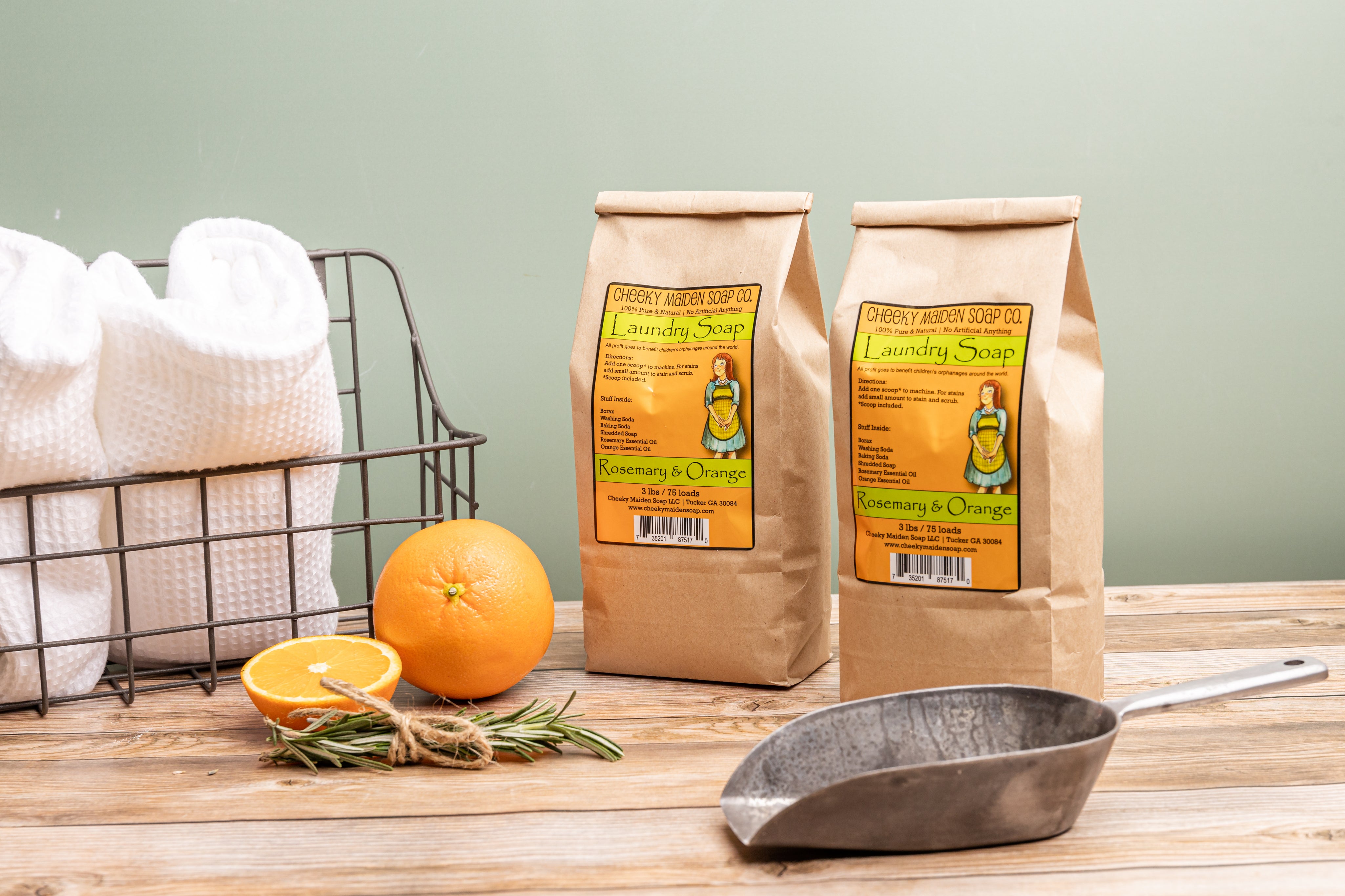 Two bags of Rosemary & Orange soap on a wooden surface with oranges and a metal scoop.