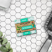Cheeky Maiden Tea Tree Shampoo bar on a tiled bathroom floor with a plant and loofah.