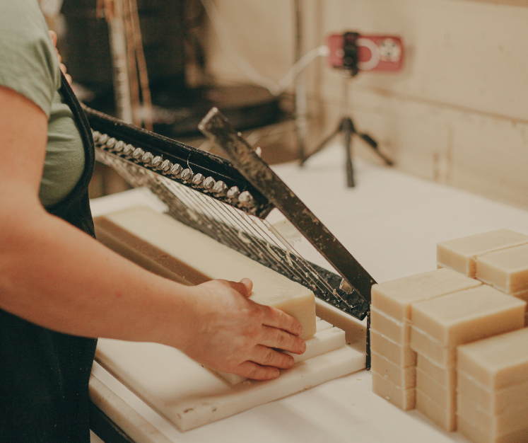 Person cutting soap bars with a large knife on a white surface, with stacked soap bars in the background.